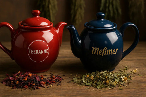 Zwei Teekannen aus Keramik – eine rote von Teekanne und eine blaue von Meßmer – stehen auf einem Holztisch, umgeben von getrocknet